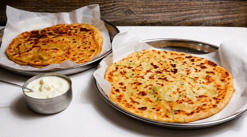 Two golden-brown parathas on metal plates with parchment paper, and a small bowl of yogurt.