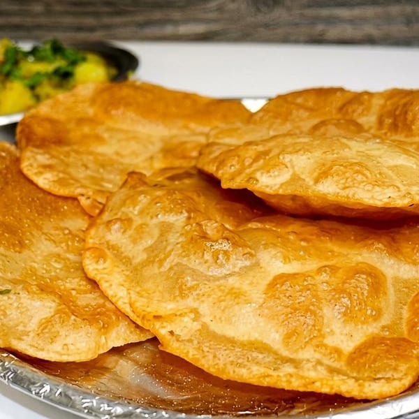 Golden-brown fried puri bread on a foil plate, with a blurred bowl of potato curry in the background.