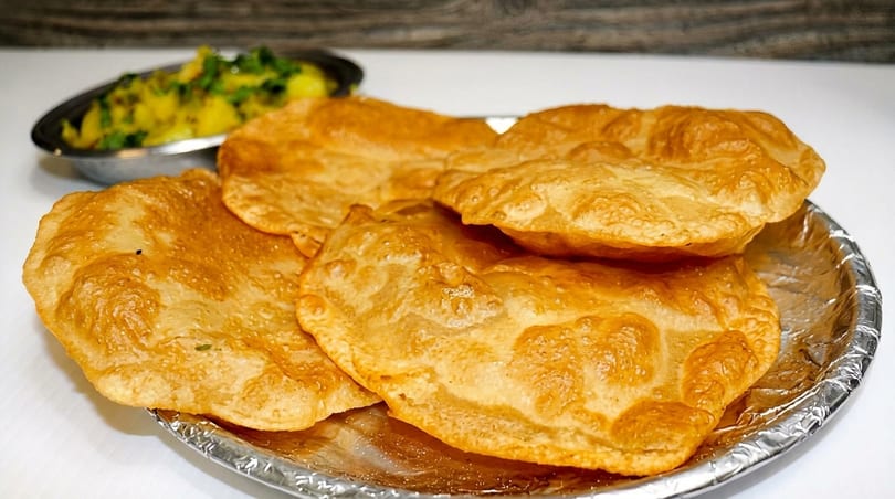 Golden-brown fried puri bread on a foil plate, with a blurred bowl of potato curry in the background.