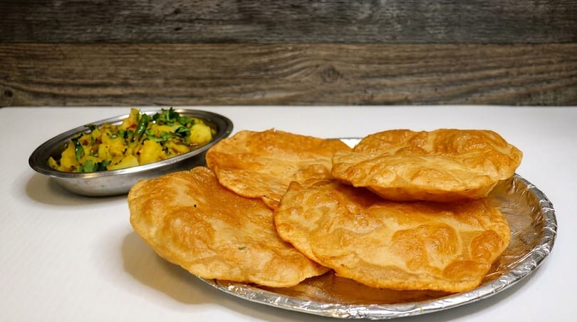 Golden fried puri bread on a foil plate with a side of potato curry.