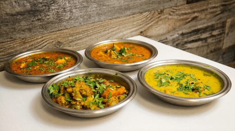 Four bowls of colorful Indian curries and stews, garnished with fresh cilantro, on a white table.