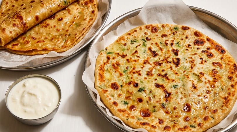 Two plates of golden-brown parathas, one folded and one round, with a small bowl of yogurt.
